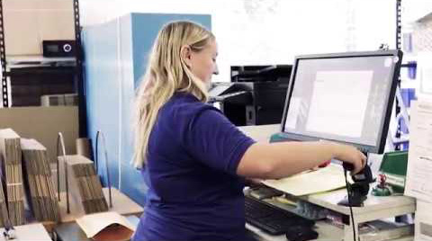 Eine Frau an einem Computer in einem Lagerraum. Sie hält einen Barcodescanner in der rechten Hand.